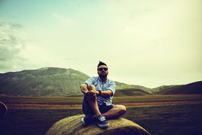 Young man sitting on hay bale against sky