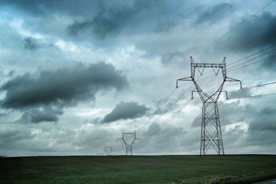 Electricity pylons on grassy field against cloudy sky