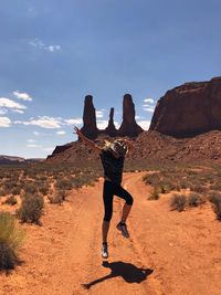 Woman jumping over dirt road against blue sky