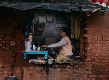Varanasi, india - february, 2018: side view of adult male in typical clothes preparing food while sitting with crossed legs on window hole of old brick building