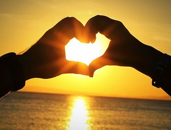 Cropped hands of people making heart shape at beach against sky during sunset