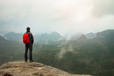 Red backpack and running suit man stands on top of a rock against background rocky foggy valley 