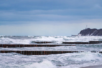 Scenic view of blue sea with foaming waves, cloudy sky and vintage long wooden breakwaters in sea