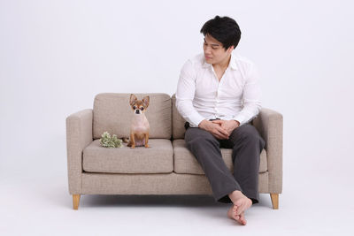 Full length of man sitting with dog on sofa against white background