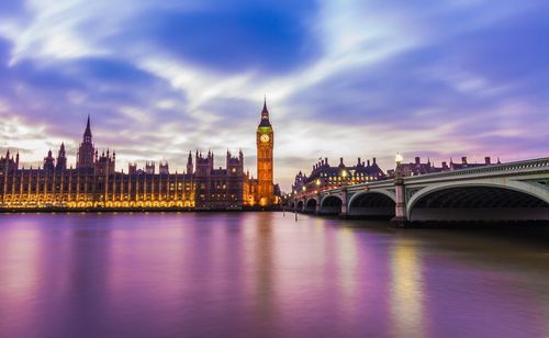 Illuminated big ben tower by parliament building and bridge against sky during sunset