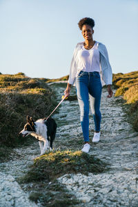 Full body of happy ethnic woman with border collie dog walking together on trail among grassy hills in sunny spring evening
