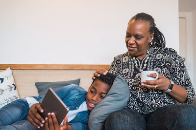 Happy boy using digital tablet while lying on sofa by grandmother at home