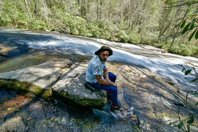 Full length of man sitting on rock in forest by a waterfall