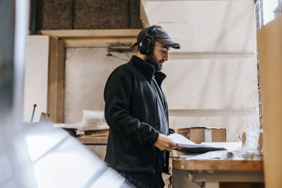 Focused male employee wearing wireless headphones and cap while reading document near table at warehouse