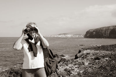 Portrait of woman looking through binoculars while standing by sea