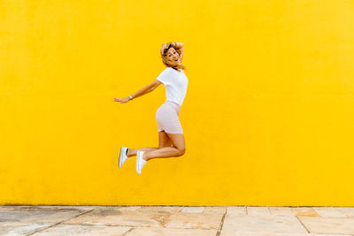 Full length of a young woman jumping against yellow wall