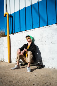 Young man wearing sunglasses sitting on skateboard against wall
