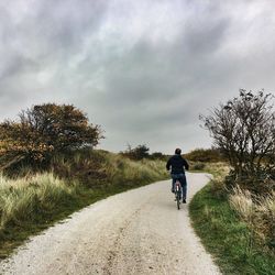 Rear view of man riding bicycle on road leading towards cloudy sky