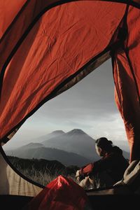 Side view of man sitting by tent against mountain and sky