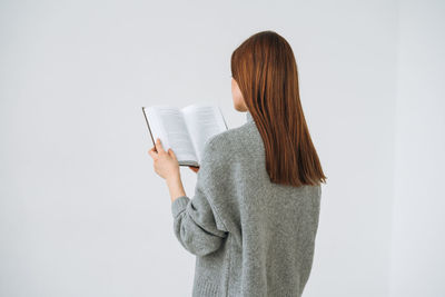 Beautiful young woman with dark long hair in grey knitted sweater reading book on white background