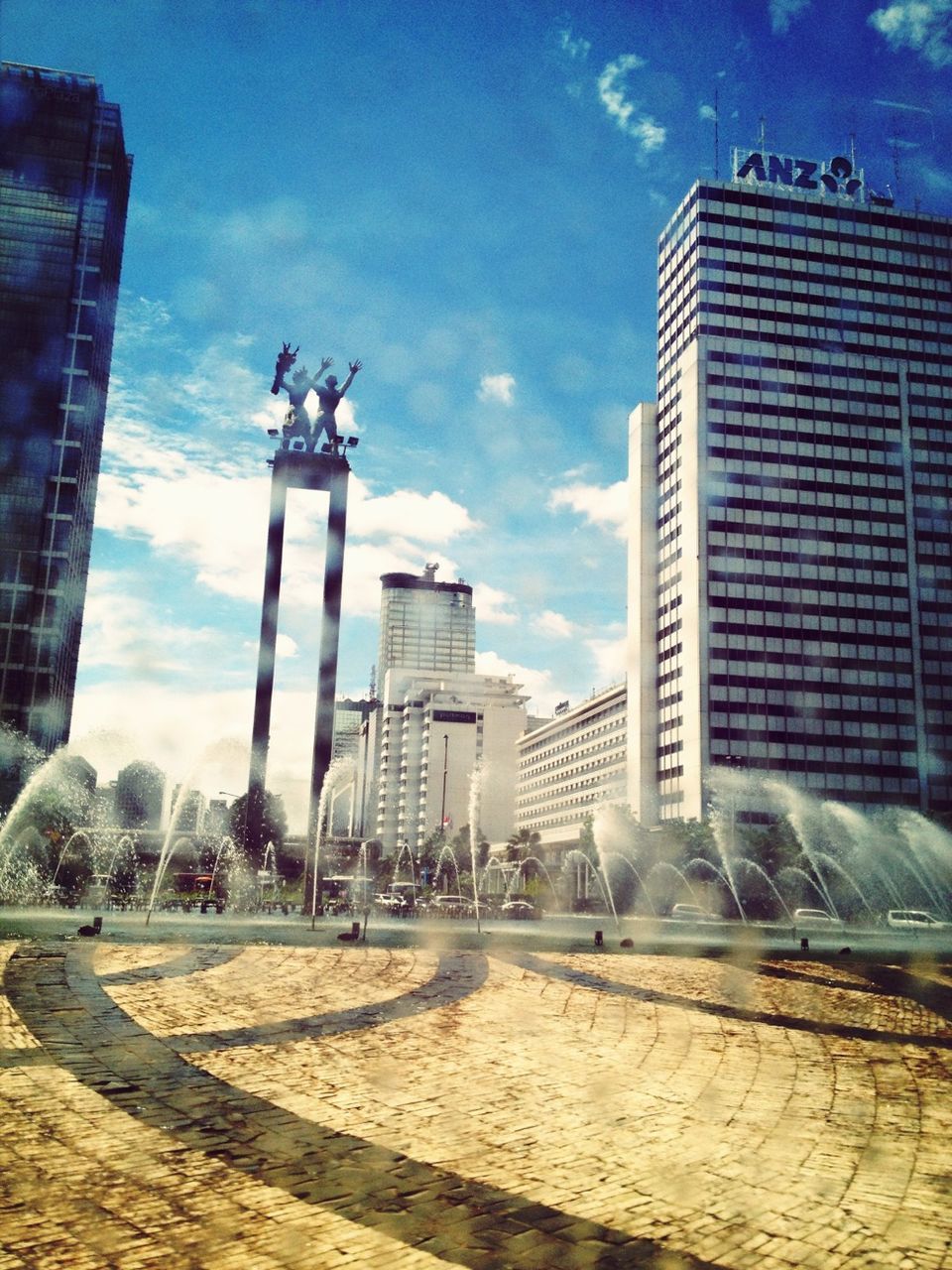 building exterior, architecture, built structure, fountain, sky, city, cloud - sky, water, sculpture, modern, building, cloud, statue, reflection, day, spraying, outdoors, sunlight, motion, glass - material