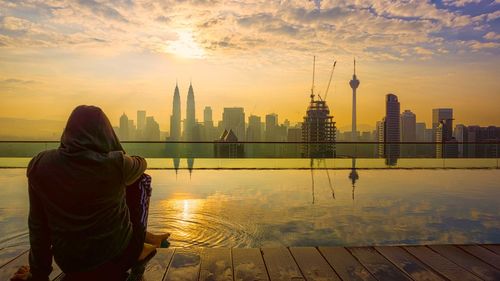 Rear view of person sitting by infinity pool with cityscape in background against sky