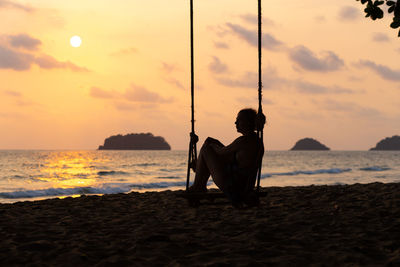 Silhouette man sitting on beach against sky during sunset