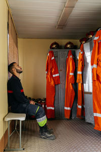 Full body side view of tired young male firefighter in uniform leaning on yellow wall against lockers with orange protective clothes hanging on doors inside fire station