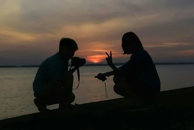 Silhouette man photographing woman gesturing peace sign at beach against sky during sunset