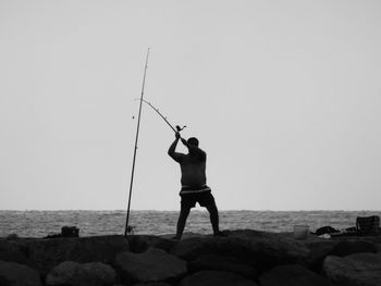 Man fishing on rocky sea shore against clear sky