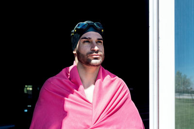 Through window view of dreamy male athlete in swimming cap and goggles with towel looking away on black background