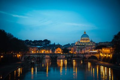 Illuminated bridge over river by buildings against sky at dusk