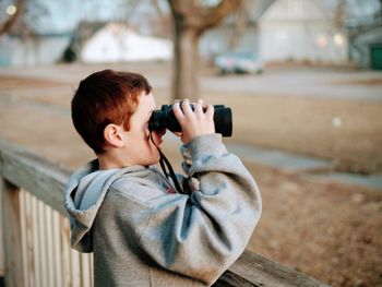 Side view of boy looking through binoculars while standing by railing