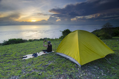 Rear view of woman sitting by sea against sky during sunset