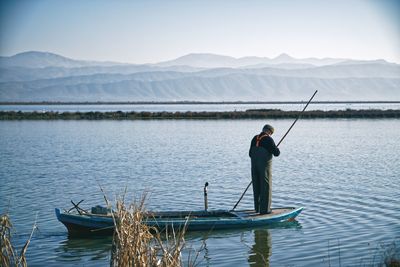 Rear view of man fishing in lake against mountains