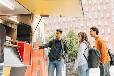 Young man pointing at menu while standing with friends by food truck in city