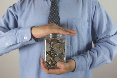 Midsection of businessman holding coins in jar against gray background