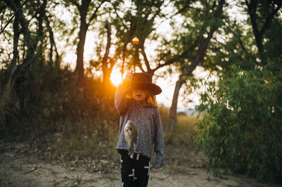 Girl wearing hat while holding fish in forest during sunset