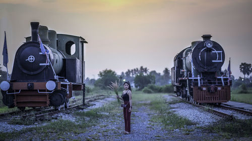 Full length of woman standing amidst train engines against sky during sunset