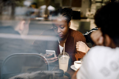 Teenage girl using mobile phone while sitting with friends at cafe seen through glass window
