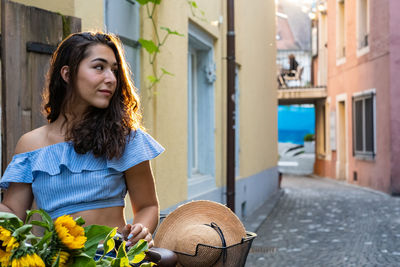 Beautiful young lady walking through an old city while pushing a bicycle with basket of sunflowers.