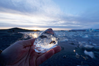 Cropped hand of person holding ice over sea against sky