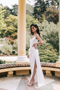 A beautiful brunette lady in an elegant wedding dress poses among the columns in the old city park