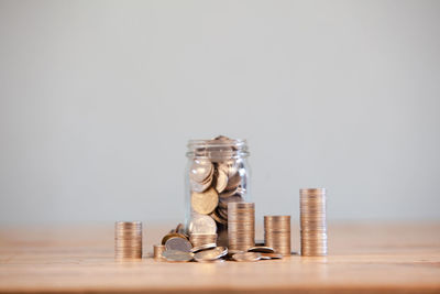 Close-up of coin stack by glass jar on table against wall