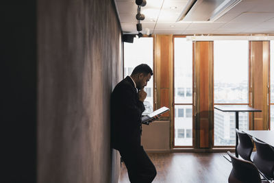 Side view of male entrepreneur with hand on chin holding digital tablet leaning against wall in board room at office