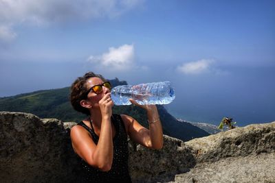 Woman drinking water from bottle against rock and sky