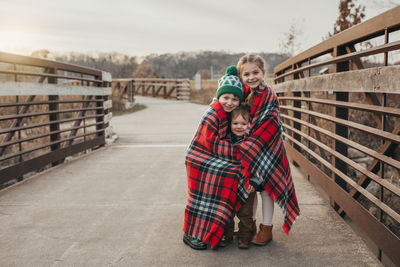 Siblings wrapped in christmas plaid blanket on bridge at sunset