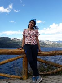 Full length portrait of young woman standing on pier over sea against mountains and sky