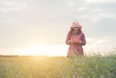 Woman with gift box standing on field against sky during sunset