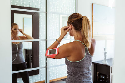 Woman in sport clothing wearing headphones while looking in mirror at home