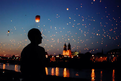 Man looking at sky lanterns flying over river during dusk