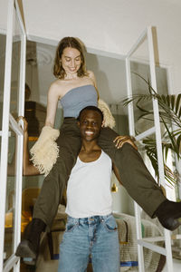 Portrait of smiling young man carrying girlfriend on shoulder at doorway of home