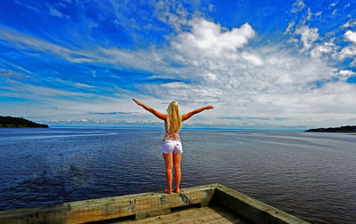 Rear view of young woman with arms outstretched standing on pier over sea against cloudy sky