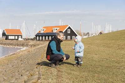 Grandfather looking at granddaughter standing on grassy field against sky during sunny day