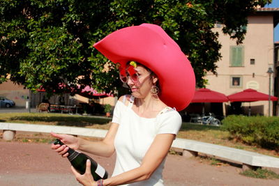 Woman holding hat while standing against trees